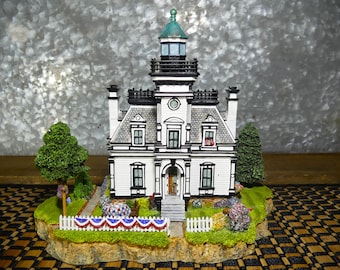 May include: A detailed miniature lighthouse with a white exterior, black trim, and a green top. The model sits on a small island with a white picket fence, greenery, and a red, white, and blue banner. The lighthouse has multiple windows and a detailed facade.
