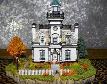 May include: A white and black Halloween-themed house with a green-topped tower, surrounded by a miniature landscape with pumpkins, a white picket fence, and autumn foliage. The house has multiple windows and a front door.