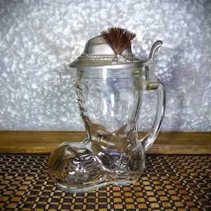 May include: A clear glass boot-shaped beer stein with a pewter lid featuring a traditional German hat with a brown feather.