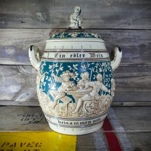 May include: A large, ceramic beer stein with a lid and handles. The stein is cream-colored with teal and brown accents. It features relief carvings of people and the text "Ein edler Wein" and "beisammen sein". A small figurine sits atop the lid.