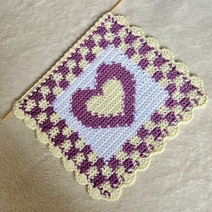 May include: A crocheted square with a heart design in the center. The heart is made of yellow yarn and is surrounded by a purple border. The square is edged with a scalloped border in yellow yarn.