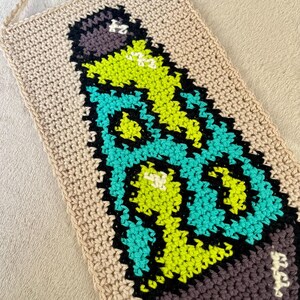 May include: Handmade crochet wall hanging featuring a colorful design. The artwork showcases a stylized bottle shape with vibrant teal, neon green, black, and purple details against a beige background. The piece is crafted with a textured stitch.