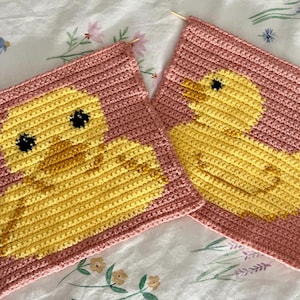 May include: Two crocheted squares with a yellow rubber duck design on a pink background. The squares are made with a textured yarn and have a simple, modern aesthetic.