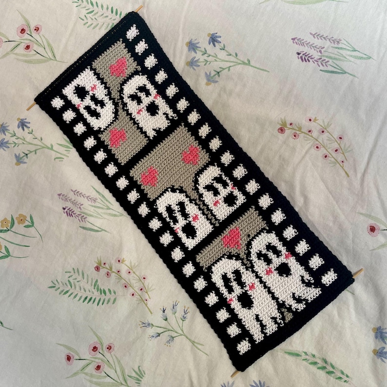 May include: A black and white crocheted wall hanging with a film strip design. Each frame features a white ghost with pink hearts.