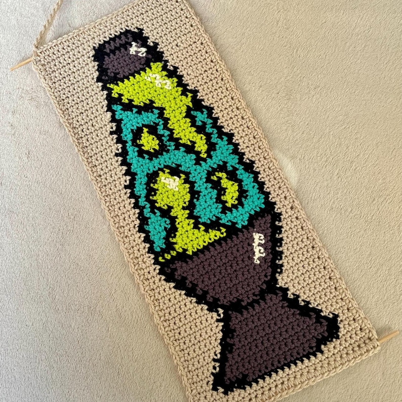 May include: Crocheted wall hanging featuring a lava lamp design. The lamp has a brown base, teal and green lava, and a black outline. The background is a beige color, and the piece has wooden dowels and a string for hanging.