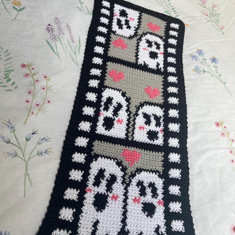 May include: A crocheted wall hanging featuring a black and white film strip design with a series of white ghosts with pink cheeks and hearts.