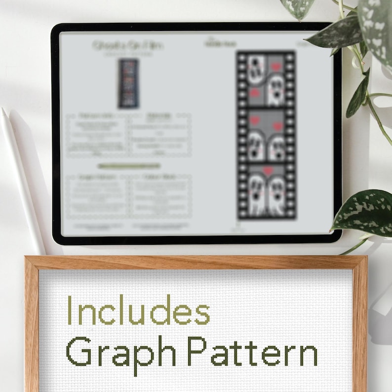 May include: A digital screen displaying a cross-stitch pattern of a film strip with three ghosts. The screen is on a wooden table with a framed piece of white fabric that says "Includes Graph Pattern".