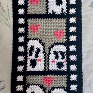 May include: A black and white crocheted wall hanging with a film strip border. The hanging features three rows of white ghosts with pink cheeks and hearts.
