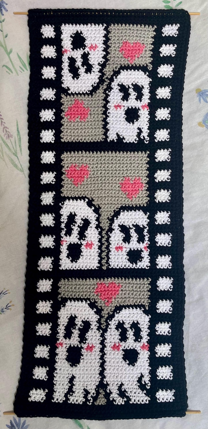 May include: A black and white crocheted wall hanging with a film strip border. The hanging features three rows of white ghosts with pink cheeks and hearts.