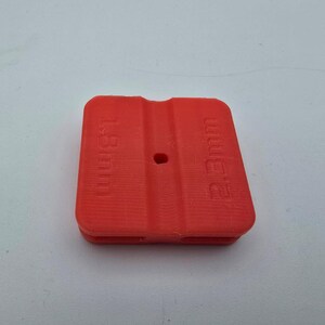 Puede incluir: Un objeto de plástico rojo brillante, de forma cuadrada, con las medidas "1.8mm" y "2.3mm" impresas. Tiene un pequeño agujero en el centro y ranuras en los bordes. Probablemente un espaciador o componente.