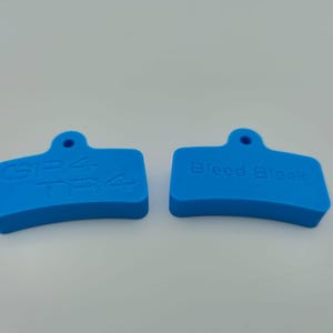 Puede incluir: Dos bloques rectangulares azules brillantes y curvos con un pequeño agujero en la parte superior. Uno tiene el texto "SRAM T4" y el otro "Bleed Block". Probablemente son herramientas o piezas para un sistema mecánico.