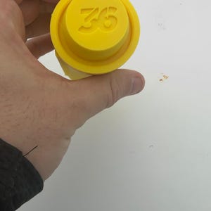 Puede incluir: Un objeto cilíndrico de color amarillo brillante con una parte superior circular elevada que presenta el número 36 en relieve. El artículo tiene una superficie lisa de color sólido y un borde distintivo alrededor de la parte superior.