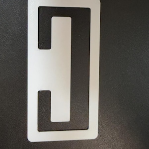 Può includere: Un oggetto rettangolare bianco con un design ritagliato unico. L'oggetto presenta una barra verticale centrale con due barre orizzontali, creando una forma a 'H' aperta. Lo sfondo è una superficie scura e strutturata.
