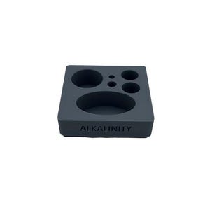 May include: A dark gray, square organizer with various circular and oval cutouts. The word "ALKALINITY" is embossed on the front. The organizer is designed to hold small items.