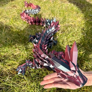 Könnte beinhalten: Eine geometrische Drachenskulptur in Rot-, Schwarz- und Grautönen. Der Drache ist aus gefaltetem Papier oder Karton gefertigt und weist scharfe, kristalline Merkmale auf. Die Skulptur wird auf grünem Gras ausgestellt.