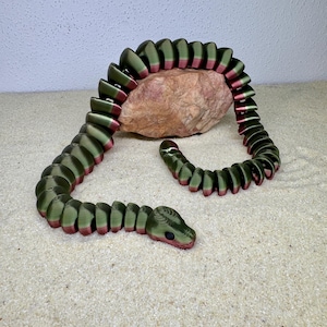 Könnte beinhalten: Eine segmentierte, grün-rote Schlangen-Skulptur aus Kunststoff. Die Schlange ist um einen Stein gewickelt und hat einen realistischen Kopf mit schwarzen Augen.