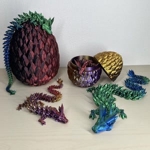 Könnte beinhalten: Fünf schillernde Origami-Drachen, von denen einer sich in einem geschlossenen schillernden Dracheneier befindet und zwei weitere in einem geöffneten schillernden Dracheneier. Die Drachen sind aus Papier gefertigt und haben eine Vielzahl von Farben, darunter Grün, Blau, Rot und Lila.