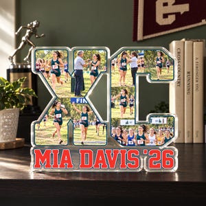 May include: A clear acrylic photo display shaped like the letters XC, featuring multiple action photographs of a cross country runner. The base of the display is printed with the red text MIA DAVIS '26 in a bold, block font style.