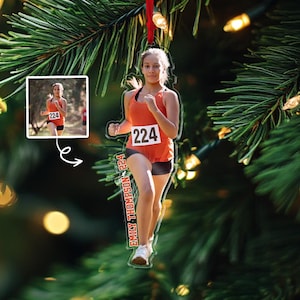 May include: A clear acrylic ornament featuring a runner in an orange top and black shorts, bib number 224. The ornament includes a smaller inset image of the runner and the name "Emily Thompson 224" printed vertically. The ornament is hanging from a red ribbon.