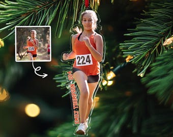Könnte beinhalten: Ein klarer Acryl-Ornament mit einer Läuferin in einem orangefarbenen Oberteil und schwarzen Shorts, mit der Startnummer 224. Das Ornament enthält ein kleineres inset Bild der Läuferin und den Namen "Emily Thompson 224" vertikal gedruckt. Aufgehängt an einem roten Band.