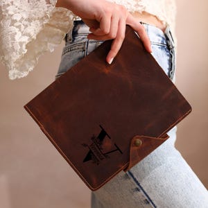 May include: A brown leather journal with a flap closure and a snap. The journal features a personalized design with the letter "V" and the name "Veronica" in black. The leather has a distressed look.