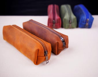 May include: Two brown leather pencil cases with zippers. The cases are lined up next to each other on a white surface. There are four other pencil cases in the background, one red, one green, and two blue.