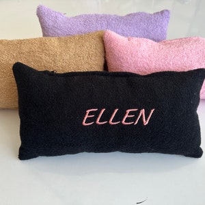 May include: A collection of rectangular, plush pillows in various pastel colors: black, tan, pink, and lavender. The black pillow has the name "ELLEN" embroidered in pink thread. The pillows are made of a soft, textured material.