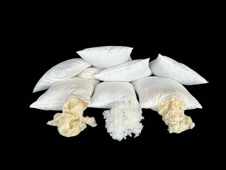 May include: A group of eight white pillowcases with different types of filling. The pillowcases are arranged in a pyramid shape with the filling spilling out of the bottom of the pillowcases. The fillings include white polyester fiberfill, white down feathers, and cream-colored wool.