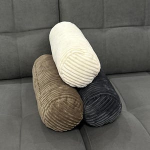 May include: Three cylindrical pillows in different colors and textures. The top pillow is white with a ribbed texture. The middle pillow is brown with a corduroy texture. The bottom pillow is dark gray with a corduroy texture.