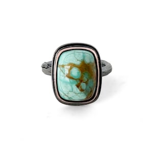 May include: A ring featuring a rectangular turquoise stone with brown matrix veining, set in a rose gold bezel and framed by a dark, oxidized silver border. The ring band has a textured, bark-like finish, providing a rugged contrast to the smooth stone.