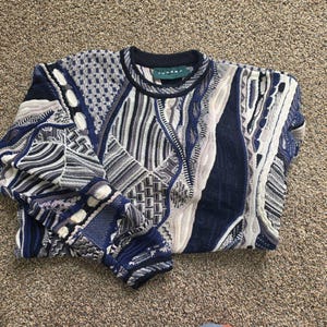 May include: A long-sleeve crew neck sweater featuring a complex, multi-textured knit pattern in shades of navy blue, white, and grey. The garment displays a variety of geometric and abstract knit designs throughout, with a Tundra brand label at the neck.