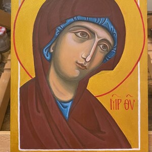 May include: A hand-painted icon of the Virgin Mary, with a gold halo and a red and blue robe. The icon is on a wooden background and has the text "MP OY" written in white.