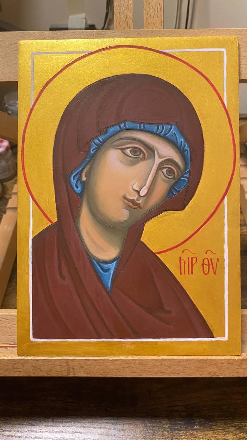 May include: A hand-painted icon of the Virgin Mary, with a gold halo and a red and blue robe. The icon is on a wooden background and has the text "MP OY" written in white.