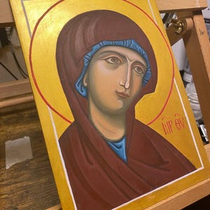 May include: A hand-painted icon depicting a woman with a blue head covering and a red robe. The icon is set against a gold background with a red halo. The text "HIP EV" is written in the lower right corner.