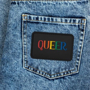 May include: A black patch with the word "QUEER" embroidered in rainbow colors is sewn onto a blue denim pocket.