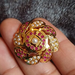Può includere: Un anello d'oro con un designo floreale. L'anello è adornato con piccole pietre rosse e bianche.