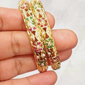 Puede incluir: Dos brazaletes dorados con diseños intrincados. Los brazaletes están adornados con piedras preciosas rojas, verdes y blancas, creando un patrón colorido. El oro tiene un acabado pulido, realzando la elegancia general de la joyería.
