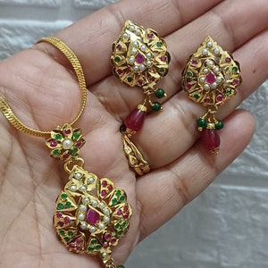 May include: A gold necklace and earrings set with a pendant and earrings featuring a leaf design with red and green stones and white pearls.