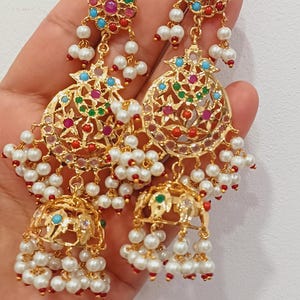 May include: A pair of gold-toned earrings with intricate filigree designs. The earrings feature colorful gemstones in red, green, blue, and pink, and are adorned with dangling white pearls. The earrings have a traditional, ornate style.