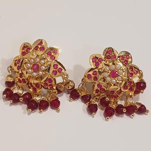 May include: A pair of gold-coloured earrings with a floral design. The earrings feature small, round, red gemstones and white pearls. Dangling from the bottom are clusters of red, round beads.