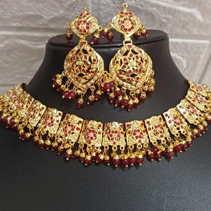 May include: A gold-toned jewelry set featuring a necklace and matching earrings. The set is adorned with red gemstones and small white accents, with dangling red beads. The earrings and necklace have an ornate, detailed design.