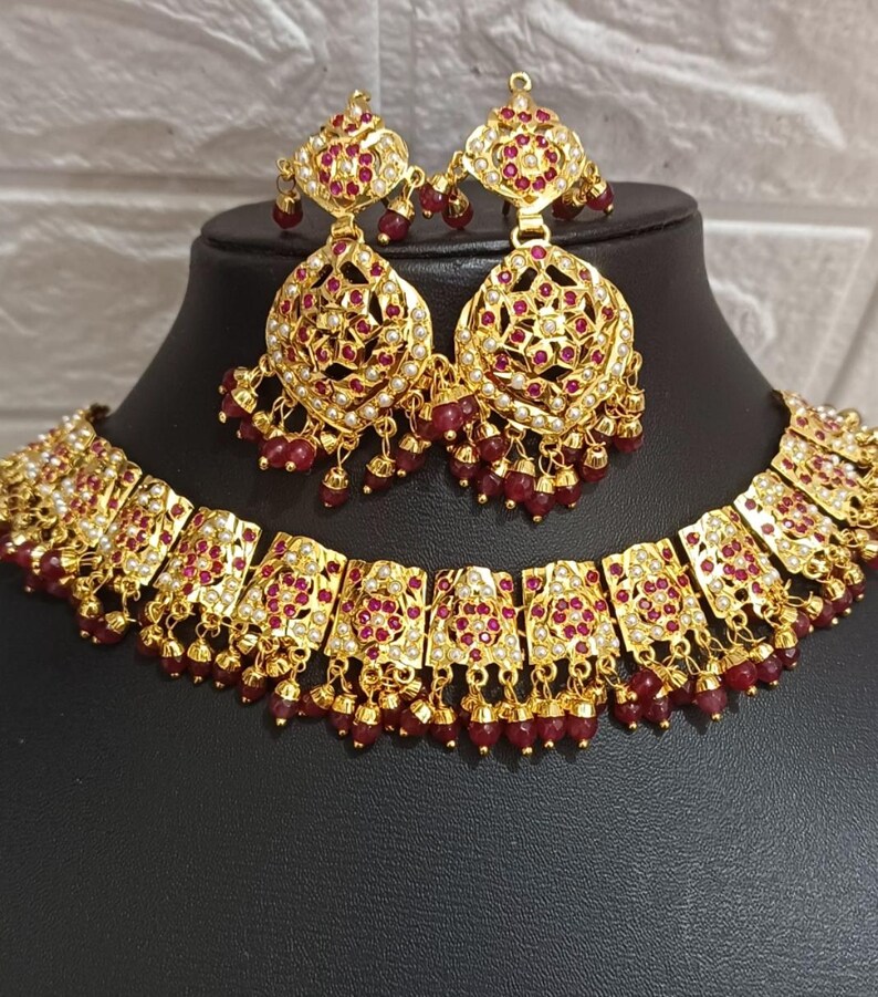 May include: A gold-toned jewelry set featuring a necklace and matching earrings. The set is adorned with red gemstones and small white accents, with dangling red beads. The earrings and necklace have an ornate, detailed design.