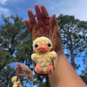 Peut inclure: Un jouet fait à la main en forme de caneton jaune en peluche, avec un bec et des pieds couleur pêche, suspendu à un cordon marron. Le caneton est dans une balançoire marron, sur fond de ciel bleu et d'arbres.