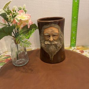 May include: A brown ceramic vase featuring a detailed relief of a bearded man's face. The cylindrical vase is on a brown surface. A small glass vase with pink and white flowers is to the left of the vase.
