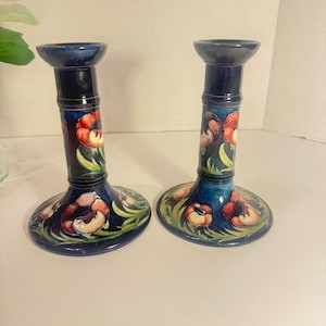May include: A pair of dark blue ceramic candle holders featuring a hand-painted floral motif with red and orange poppy-like flowers and green foliage. Each candlestick has a wide circular base, a cylindrical stem, and a flared top rim.