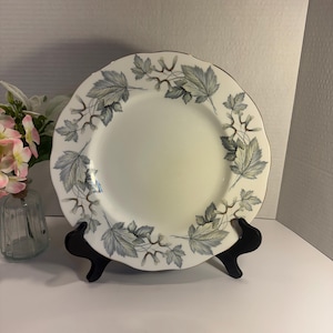 May include: A round ceramic dinner plate with a scalloped edge and a gold-toned rim. The plate features a white centre and a border decorated with a repeating pattern of grey and blue maple leaves and winged seeds arranged in a circular design.