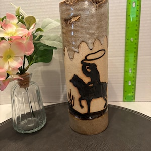May include: A ceramic vase featuring a cowboy riding a horse design. The vase is brown and white with a textured glaze.