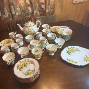 Peut inclure: Un ensemble de service à thé vintage avec une théière blanche, des tasses, des soucoupes et des assiettes. L'ensemble est orné de roses jaunes et de bordures dorées. L'ensemble comprend une théière, des tasses, des soucoupes et des assiettes.