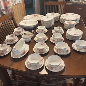 Peut inclure: Un ensemble de service à thé vintage avec un motif de roses. L'ensemble comprend des tasses, des soucoupes, des assiettes, un plat de service, une crémière et un sucrier. La porcelaine est blanche avec des motifs de roses roses et verts.