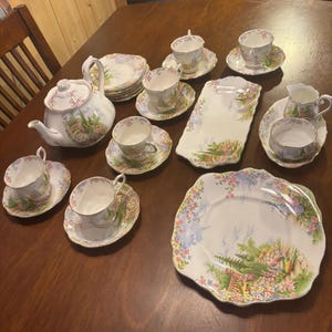 May include: A porcelain tea set featuring a white base with colourful floral garden scenes and scalloped edges. The set includes a teapot with a lid, several teacups with matching saucers, a rectangular serving tray, a creamer, and a large square plate.